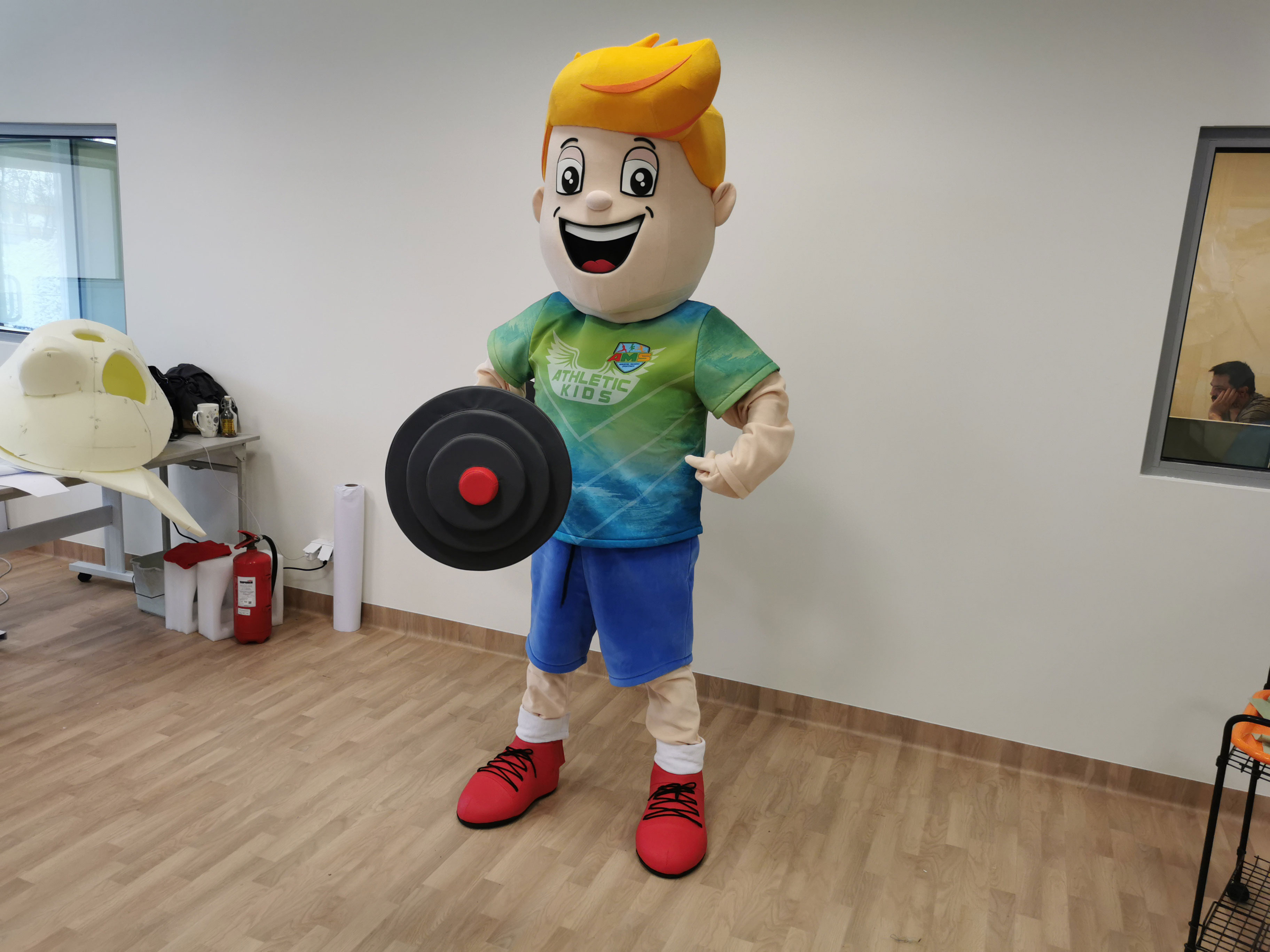 Na fotografii widnieje maskotka w postaci sztangisty pozująca do zdjęcia w siedzibie firmy Elabika